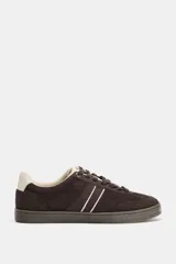 Zapatillas deportivas de gamuza en color marrón oscuro, con detalles de franjas laterales en color crema y refuerzo en el talón. Presentan cierre mediante cordones y suela de goma texturizada.