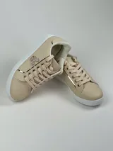 Zapatillas deportivas de cuero sintético en color blanco, beige y negro, con cordones y aplique decorativo dorado con forma de moño.