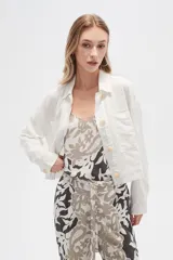 Chaqueta cropped de manga larga, color blanco con finas rayas verticales en tono claro. Presenta cuello camisa, cierre frontal con botones de madera y dos bolsillos delanteros con tapa a la altura del pecho.