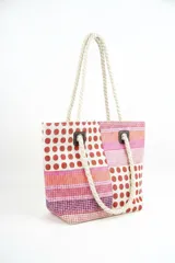 Bolso tipo tote con diseño de paneles en bloques que combinan estampado de lunares rojos sobre fondo blanco y franjas horizontales en tonos rosados, naranjas y morados. Cuenta con asas de soga gruesa de algodón en color crudo, sujetas con ojales metálicos y refuerzos de cuerina marrón.