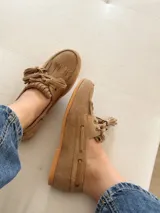 Mocasín tipo náutico de gamuza color camel, con detalle de flecos y borlas en el empeine, cordones laterales y suela plana de goma.