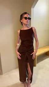 Vestido largo de color marrón con diseño de escote recto y tirantes finos. Presenta un detalle de nudo frontal en la cintura que crea un efecto drapeado y una abertura central pronunciada que deja ver una capa inferior más corta.