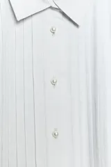 Camisa de cuello solapa y manga larga con puños anchos. Presenta un diseño con pliegues verticales en el frente y cierre frontal con botones tipo perla.