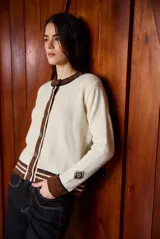 Cardigan clásico de largo medio color crudo, de manga larga, con cuello redondo, puños y ribetes en marrón con rayas blancas, y cierre frontal de botones marrones.