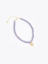 Collar de cuentas de vidrio color lila, con un charm dorado en forma de llama en el centro. Cuenta con cadena de extensión y cierre de mosquetón dorados.