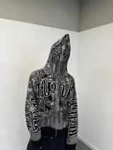 Buzo con cierre frontal completo, confeccionado en tejido de punto grueso de algodón. Presenta un diseño estampado integral con motivos gráficos y tipografía estilo gótico en tonos gris y negro, capucha integrada, puños y ruedo acanalados en color negro sólido.