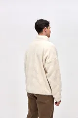 Campera de sherpa con diseño capitoneado en color crema. Presenta cierre frontal completo, cuello alto, bolsillos laterales y un bolsillo en el pecho con cierre en contraste. Incluye detalles en nylon y bordado decorativo en el pecho.
