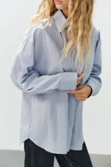 Camisa oversize de manga larga con cuello solapa, cierre frontal de botones y bolsillo de parche en el pecho, con diseño de rayas verticales celestes y blancas.