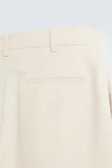 Pantalón de corte recto, color beige claro, con pliegues frontales y bolsillos de vivo en la parte trasera.