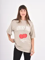 Remera de corte oversize color beige, con cuello redondo y mangas cortas. Presenta un estampado frontal con la frase 'Amore Mio', un dibujo de dos cerezas y la leyenda 'South Tuscany Italy'.
