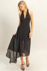 Vestido largo negro con escote halter y espalda descubierta. Presenta un diseño asimétrico con una capa superpuesta de tela semitransparente que cae lateralmente, creando un efecto de movimiento y elegancia.
