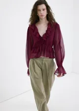 Blusa bordó de tejido fluido y semi transparente, con escote en V pronunciado, mangas largas abullonadas con volantes en los puños y detalles de volantes en el cuello y el ruedo.