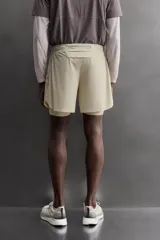 Short de running 2 en 1 color beige, confeccionado en tejido técnico ligero y elástico con malla interior integrada. Presenta cintura elástica ajustable con cordón, bolsillo trasero con cierre y aberturas laterales en el bajo.