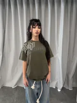 Remera de algodón color verde oliva con estampado gráfico en serigrafía plastisol. Presenta un diseño con caracteres orientales en blanco y tipografía cursiva en gris, con cuello reforzado y grifa de la marca en la manga.