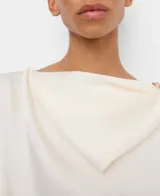 Top fluido satinado color marfil, con línea rectangular, cuello pañuelo que gira desde el delantero hacia la espalda sujeto con hebilla decorativa dorada en el hombro izquierdo y manga larga con dos botones en el puño.