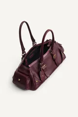 Bolso de hombro alargado de piel color borgoña con doble asa, detalles y hebillas metálicas doradas, charm de espejo colgante y bolsillos laterales exteriores.