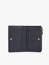 Billetera bifold Tommy Hilfiger de diseño compacto en color gris. Presenta un cierre superior con cremallera y un detalle frontal decorativo con broche metálico que incluye el logo de la marca. Cuenta con múltiples compartimentos internos para tarjetas y billetes.