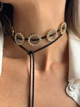 Choker negro con cuatro dijes redondos dorados, cada uno con un centro de resina transparente y borde de strass. Se ajusta con cordones negros.