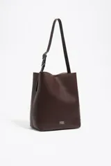 Bolso de hombro grande de estructura rígida, color marrón oscuro, confeccionado en piel vacuna. Presenta un cierre de cordón y un asa ajustable con hebilla metálica. El logo metálico de BIMBA Y LOLA es visible en la parte inferior frontal.