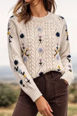 Sweater de lana merino color beige, con tejido de punto trenzado y bordados artesanales de flores en tonos azules, amarillos y morados, cuello redondo y mangas largas.