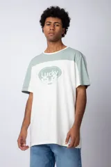 Remera de corte holgado con diseño color block, combinando paneles en color verde menta y blanco. Presenta un estampado frontal con la palabra 'Lucky' dentro de un óvalo y dibujos de flores con lentes de sol.