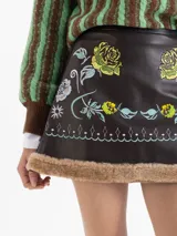 Minifalda de cuero ovino color marrón oscuro con bordados florales en tonos amarillos y verdes. Presenta un ruedo con detalle de piel sintética afelpada y una estructura definida.