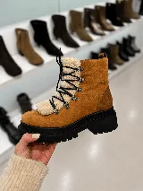 Bota tipo borcego de cuero color marrón, con suela track de goma negra. Presenta un aplique frontal desmontable de textil tipo corderito en color crema, con cordones negros y ganchos metálicos.