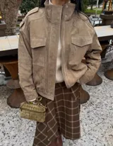 Chaqueta de gamuza en color marrón oscuro, con cuello alto, cierre frontal oculto y bolsillos tipo cargo en el pecho. Presenta un diseño de corte holgado con detalles de charreteras en los hombros.