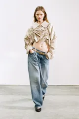 Chaqueta corta color beige, confeccionada en gabardina, con cuello solapa redondeado, mangas largas con frunces y detalle de frunces con lazada en el delantero, cerrando con botones.