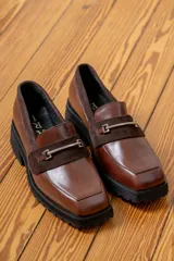 Mocasín de cuero vacuno color marrón, con suela track de goma negra, puntera cuadrada y detalle de herraje metálico sobre una banda de gamuza en el empeine.