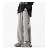 Pantalón deportivo gris claro, estilo cargo o jogging, con franjas negras verticales en los laterales de las piernas. Posee tobillos regulables y cintura ajustable.