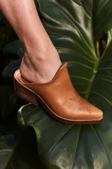 Mule de cuero color suela con diseño de punta fina y bordado decorativo en el empeine. Presenta un taco bajo de madera y una silueta abierta en el talón.