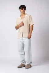 Camisa de punto calado en color beige, de manga corta y corte relajado. Presenta cuello tipo bowling, cierre frontal con botones al tono y una estructura liviana y respirable confeccionada en algodón.