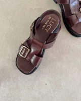 Sandalias de cuero color marrón con suela de goma negra, tiras anchas y hebilla metálica.