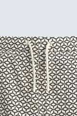 Bañador corto de hombre con cintura elástica ajustable mediante cordón blanco. Presenta un estampado geométrico repetitivo en tonos oscuros (negro/marrón oscuro) y blanco/beige. Incluye bolsillos laterales y un bolsillo trasero de vivo.