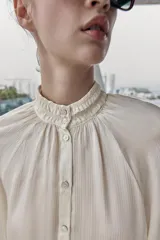 Blusa fluida de color blanco roto o crema, con cuello alto fruncido y manga larga con puños elásticos tipo nido de abeja. Presenta un frente abotonado con botones pequeños y brillantes.