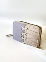 Billetera pequeña de cuero color lila y beige con cierre metálico y detalle de cordones cruzados en el frente.