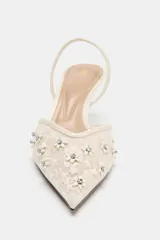 Zapato de tacón destalonado en color crudo, con cuerpo de mesh transparente con bordados florales y aplicación de abalorios brillantes. Cuenta con tira trasera elástica, tacón medio fino y acabado en punta.