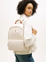 Mochila Tommy Hilfiger de nailon color beige, con diseño curvo, bolsillo frontal con cierre y detalles en cuero sintético marrón en el asa superior. Cuenta con el monograma TH metálico dorado en la parte delantera y correas ajustables.