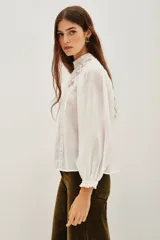 Camisa blanca de estilo romántico con cuello alto, confeccionada en algodón con detalles de encaje y bordados en el frente. Presenta mangas largas abullonadas con puños elásticos y terminaciones de encaje.