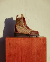 Botas de caña media con diseño de cuadros escoceses en tweed, combinadas con detalles en cuero marrón. Presentan cierre con cordones, lengüeta con logo grabado y suela de goma robusta con plataforma dentada.
