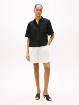 Camisa de manga corta de corte amplio confeccionada en puro lino, de color negro, con cuello inglés y un bolsillo de parche en el pecho que incluye un sutil logo bordado. Presenta hombros ligeramente caídos y cierre frontal de botones.
