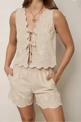 Conjunto color beige compuesto por chaleco sin mangas con cierre frontal de lazos y detalles bordados florales, y short a juego con bolsillos laterales y cintura elástica.