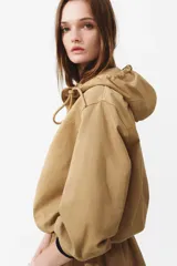 Campera popover color tostado confeccionada en sarga de mezcla de algodón y lyocell. Cuenta con capucha con cordón ajustable, medio cierre frontal y un amplio bolsillo delantero con solapa. Presenta diseño de hombros caídos, canesú frontal, mangas abullonadas con pliegues, puños con cierre ajustable de botón y bajo con cordón interior para fruncir.