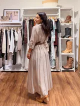 Vestido largo de seda en color beige, con mangas tres cuartos, cuello camisero y falda amplia con caída fluida. Incluye un cinturón tejido en tono marrón que marca la cintura.
