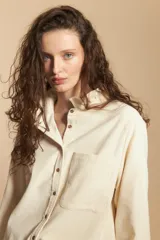Camisa de pana color crema con cuello camisero con volados, cierre frontal de botones y mangas largas con puños abotonados. Presenta un bolsillo aplicado en el pecho y una silueta relajada.