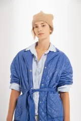 Campera azul capitoneada de nylon con forro de algodón y relleno de poliéster. Tiene ruedo curvo, largo midi, cinta para atar a la cintura y bolsillos delanteros.