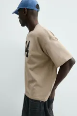 Sudadera de manga corta color beige, corte relajado (relaxed fit) con cuello redondo y número "74" bordado en negro en el frente.