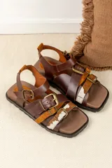 Sandalias de cuero marrón con diseño de tiras múltiples, hebillas metálicas ajustables y detalle de cuero con pelo estampado en la parte frontal. Presentan una base cuadrada y cierre de pulsera en el tobillo.