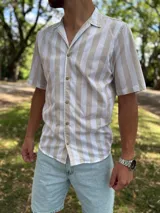 Camisa de manga corta con estampado de rayas verticales en tonos beige y blanco, cuello abierto tipo cubano y cierre frontal con botones marrones.